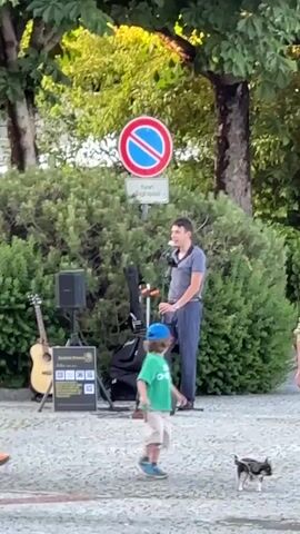 Here is a description of the video:

The video shows a busker performing on a paved street with a microphone and amplifier. A sign with a red circle with a blue background and a white bar through the middle, along with text that reads "Fuori dagli spazi," is above the bushes behind him.  A man with sunglasses, a t-shirt, and jean shorts walks by, and a young child in a green shirt and baseball cap runs up and gives him a high-five. They briefly pose and the man puts his arm around the child as they look towards the singer. A dog follows the child in the street. A guitar is on the ground next to the speaker. Several trees surround the area.