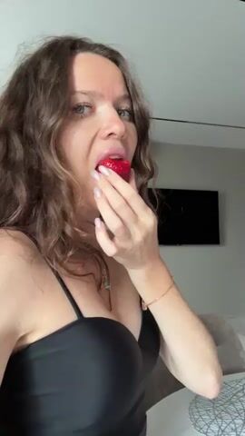A woman with long, wavy brown hair is shown in a close-up shot, wearing a black spaghetti strap top. She is holding a strawberry to her mouth with her right hand, her fingers touching her lips. Her eyes are looking upwards and slightly to the left, and her lips are pursed as if she is about to eat or has just eaten the strawberry. Her left hand is held up, palm facing outwards, near her face. The background is slightly out of focus, but appears to be an indoor setting with a wall and what looks like a television screen mounted on it. The lighting is soft, illuminating the woman's face and upper body.