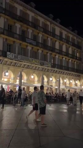 The video shows a nighttime scene in a European city square. The square is lined with buildings featuring arcades at ground level. Several people are visible walking and sitting at outdoor cafes under the arcades. Some tables are occupied, and umbrellas are set up for shade. The buildings have multiple stories with balconies and numerous windows, some illuminated. A flag, appearing to be Greek, is visible on the facade of one building. The ground is paved, and the lighting from the cafes and lampposts casts reflections on the wet pavement, indicating it may have recently rained. The atmosphere appears lively, with activity at the cafes.