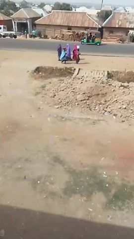 The video shows a dirt lot with piles of debris next to a paved road in what looks to be a rural or developing area. Four individuals wearing full-coverage clothing, including face coverings, are walking alongside the road, away from the camera. They are wearing purple, light blue, pink, and dark colors. One person appears to be carrying something on their head. In the background, there is a motorcycle on the road and buildings with corrugated metal roofs and stacks of firewood. The ground is dry with sparse vegetation. The video is taken from an elevated position, possibly a window.