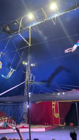 A trapeze artist in a blue and white costume swings from a trapeze bar in a circus tent. The tent has a blue fabric ceiling with spotlights. Below the trapeze, there is a red curtain backdrop with gold trim. The audience is seated in the lower portion of the frame. Another trapeze artist is visible on a platform to the left, and their shadow is cast on the tent ceiling. The video captures the trapeze artist mid-swing.