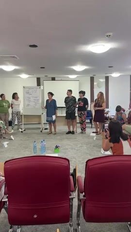 A group of people are in a room. Some are standing in a semi-circle facing a whiteboard. One person is standing by the whiteboard and appears to be presenting. Others are seated in chairs arranged in a circle. There are water bottles and some markers on the floor. The room is brightly lit with fluorescent lights on the ceiling.