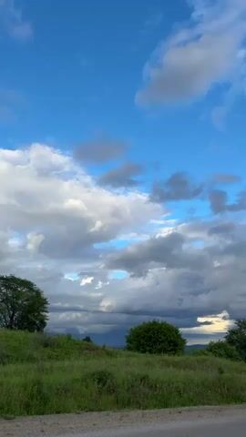 This is a vertical video of a rural landscape in Teployozersk, Russia. The scene is set under a dynamic sky with a mix of bright blue patches and dramatic, heavy grey clouds, suggesting variable weather. The sun appears to be breaking through or setting, casting a warm light on some of the clouds and distant hills.

The foreground features a grassy, unpaved shoulder next to a paved road that curves to the right. Lush green grass and vegetation cover the rolling hills that dominate the midground. Scattered trees and a few small houses with dark roofs are visible nestled amongst the hills. The background consists of dark, green, and tree-covered mountains stretching across the horizon.

The overall mood is one of natural beauty and tranquility, possibly with a hint of impending weather. The time of day appears to be late afternoon or early evening, given the quality of the light. There are no people or specific objects of interest clearly discernible, making it a landscape-focused video. The video captures the vastness of the sky and the gentle undulation of the Russian countryside.