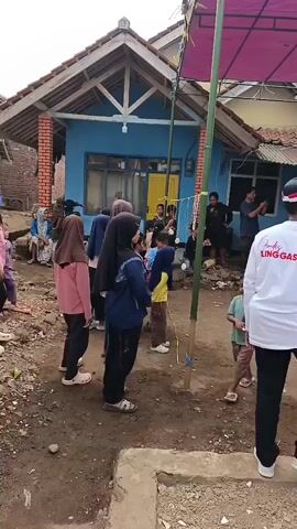 A group of children and adults are gathered in a dirt yard in front of a blue house. A young boy in a light green shirt and brown pants stands near a bamboo pole with a string tied to it. A group of girls in hijabs and colorful shirts are watching him. Two men stand near the house, one smoking a cigarette. Another man in a white shirt with the words "LINGGASIRNA" printed on the back stands facing away from the camera. A pink canopy is visible overhead. The scene appears to be some kind of outdoor event or celebration.