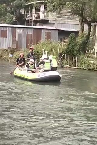 A yellow and white inflatable raft with "AYU ADVENTURE" written on it is moving down a river. There are five people in the raft, all wearing life vests and helmets. Two people in the front are rowing with paddles. The other three are sitting behind them. The raft is passing a residential area with houses on stilts and corrugated metal roofs. The river is murky and flowing. The scene appears to be a recreational activity, possibly whitewater rafting.
