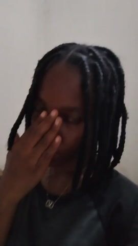 A close-up video captures a young person with dark skin and braided hair. They are wearing a dark-colored shirt and a necklace with a pendant. The person initially has their hand covering part of their face, looking towards the camera. They then move their hand away, revealing their full face and smile before raising their other hand in a waving gesture. The background appears to be a plain, light-colored wall with some stacked items on the left side. The lighting is soft and even.