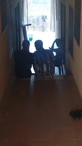 A hallway, tiled in brown, leads to a brighter outdoor space. Three people are positioned at the end of the hallway, looking outward. The person on the left is wearing a dark-colored shirt and sits on the floor. The person in the middle is also seated on the floor and is wearing a colorful patterned shirt. The person on the right is seated on a plastic chair. A small plastic step ladder and a light green bucket are visible in the bright area beyond the hallway. A pair of dark-colored sandals is on the floor near the camera.