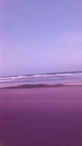 The video shows a view from inside a moving vehicle, focusing on a beach scene. The sky is a pale lavender color. A sandy beach meets the ocean, where small waves can be seen. A purple-toned road runs parallel to the beach. The camera is moving, creating a blurring effect. No people are visible in the frame. The overall color tone of the video is purple.