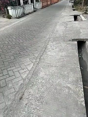 This is a low-angle, handheld video taken from a street-level perspective. The road is paved with interlocking bricks and concrete. On the left side of the road are buildings with white pillars and a fence. A pink flowering plant is visible in front of one building. There are flags on poles. On the right side of the road, there is a concrete drainage ditch with grates. The road stretches into the distance, lined with more buildings and trees. A person on a motorcycle is traveling down the road in the distance. The overall environment appears to be a residential street in a rural or semi-rural area. The lighting suggests it is daytime, possibly late afternoon.