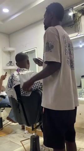 The video shows a man in a light-colored t-shirt with black shorts giving another man a haircut in what appears to be a barber shop. The barber is standing behind the seated client, using an electric razor on the client's hair. The t-shirt of the barber has a graffiti-style design on the back that reads "WE BREAK THE RULES WITH HEART." A woman wearing a headscarf sits in the background, looking at her phone. There is a mirror on the wall behind the barber. The floor is tiled and there's a small bicycle decoration on a shelf near the ceiling. The shop seems small, with some clutter visible. A fan is visible near the ceiling.