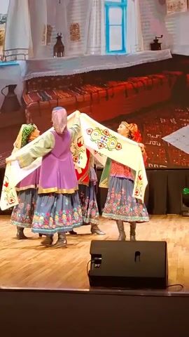 This video captures a traditional dance performance on a wooden stage. Four women in colorful, ethnic attire are participating. Their outfits include long, patterned skirts, vests worn over blouses, and headscarves. Two of the women are holding a large, decorative white cloth with embroidered colorful floral patterns, which they are manipulating in unison. The dancers are moving rhythmically, suggesting a choreographed routine. The background features a projected backdrop depicting a stylized interior scene with furniture and textiles, possibly representing a traditional home. There is a black speaker visible in the foreground on the stage. The lighting appears to be theatrical, illuminating the performers. The overall atmosphere suggests a cultural event or festival.