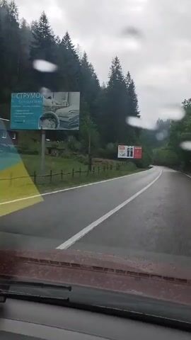 This is a first-person perspective video taken from inside a vehicle traveling on a wet, paved road. The road appears to be in a mountainous or forested area, with dense green trees lining both sides. The sky is overcast and a muted blue-gray, suggesting rain or impending rain, as indicated by the water droplets on the windshield. The road has white lane markings. A billboard is visible on the left side of the road with red and white elements, possibly advertising. There is a wooden fence and some vegetation on the left as well. The vehicle's dashboard is partially visible in the foreground. The camera is positioned to look forward through the windshield.