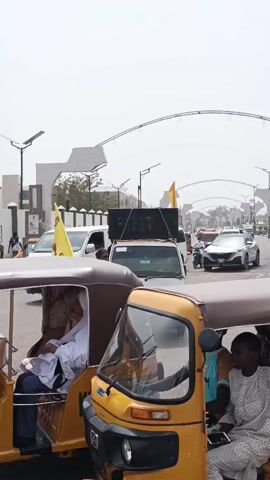 The image depicts a lively street scene in Tarauni, Nigeria, likely during a political rally or procession. In the foreground, the side of a brown, three-wheeled vehicle, possibly a Keke Napep or auto-rickshaw, is visible, with several passengers inside.

The central subject is a man of medium build, wearing a blue and white jersey with the number "29" on it, plaid shorts, and flip-flops. He has a yellow bandana or face covering around his neck and is holding a tall pole from which a yellow triangular flag unfurls. The flag prominently displays the text "APC SUPPORT GROUPS".

Behind the man stands a white HiJet van, which has a large, black, box-like speaker system mounted on its roof, suggesting music or public announcements. The van's Nigerian license plate, "NNG 834XA", is visible.

The background shows a busy street with other vehicles, including a white SUV (possibly a Nissan), another yellow three-wheeled vehicle, and a motorcycle. Distinctive modern architectural structures resembling archways or gates, along with streetlights equipped with solar panels, line the road in the distance. The sky is bright and hazy, indicating a sunny, possibly hot, midday or early afternoon. The overall scene suggests an active public gathering or political event with a sense of movement and public engagement.