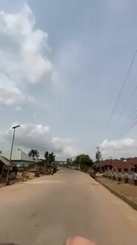 The video shows a paved road in a rural area, with buildings and vegetation along the sides. Several people are visible on the left side of the road, along with a couple of parked motorcycles. Power lines run along the right side of the road. The sky is partially covered with clouds. The camera appears to be moving forward along the road.