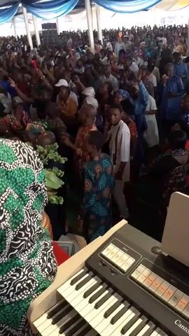 The video depicts a lively indoor event taking place in Kubwa, Nigeria, within a large tent or hall decorated with white, blue, and yellow fabric drapes across the ceiling. The floor is covered with a red carpet.

A substantial crowd of people, including adults and children, are gathered, many standing and actively participating. In the foreground, an electronic keyboard is visible, suggesting live musical performance. Several individuals, possibly choir members or performers, are dressed in matching green and black patterned traditional attire, singing and dancing energetically.

The camera often pans from behind individuals in the crowd or near the performance area, showing the packed audience. Many attendees are seen with their hands raised, indicating active engagement in worship, singing, or celebration. One person in a red long-sleeved shirt wears a green baseball cap and has the text "iArise & Prosper" printed on the back of their shirt. The atmosphere is festive and communal, with the crowd appearing to be fully immersed in the event.