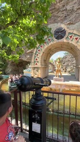 The video shows a water ride at an amusement park. In the foreground, a person is operating a cannon that appears to be part of the ride's attraction. The cannon is aimed at a boat filled with people on the water. The boat is moving towards an archway decorated with ancient-looking motifs. Behind the archway, there are statues and rock formations that create an exotic atmosphere. The surrounding environment is lush with green foliage, suggesting a warm, sunny day. The overall scene depicts a playful and adventurous experience typical of an amusement park.