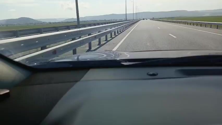 The video shows a first-person perspective from inside a car driving on a highway. The view is from behind the dashboard and windshield. The highway has multiple lanes separated by white lines. On the left side of the highway, there is a metal guardrail and beyond that, a grassy field and distant hills. On the right side, there is also a guardrail and a vast, flat field, possibly farmland, stretching to the horizon. The sky is blue with some white clouds, indicating daytime. Streetlights are visible along the side of the highway. The car appears to be in motion, as suggested by the slight blur and the receding perspective of the road. No other vehicles are prominently visible in the immediate vicinity, but there are a few in the distance. The interior of the car is partially visible, including the dashboard and part of the steering wheel area. The lighting suggests a sunny day.