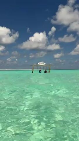 The video shows a shallow, turquoise-colored sea under a blue sky with scattered white clouds. In the distance, the sea meets the horizon. In the middle of the frame is a wooden structure with a sign reading "I [heart] SUJU." Two people are sitting in the water beneath the structure. The water's surface shimmers and appears very clear. The camera remains stationary.