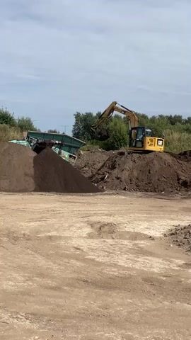 A yellow excavator is positioned on top of a dirt mound. In front of the excavator is a green conveyor belt machine with material piled next to it. The excavator's arm is extended, and its bucket is scooping material from the conveyor belt machine and depositing it into a large, dark pile of dirt. The environment appears to be an outdoor industrial or construction site, with more dirt piles and trees in the background under a cloudy sky.