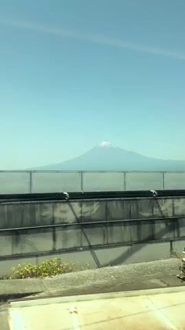 The video, shot from the interior of a moving train, captures a glimpse of Mount Fuji on a clear, bright day. The train car's window and interior are visible in the foreground. As the train moves, the landscape unfolds, revealing houses, fields, and a commercial building at the base of Mount Fuji. The mountain itself is partially obscured by a slight haze, but its iconic snow-capped peak is clearly visible against the clear blue sky. The train continues on its journey, eventually obscuring the mountain from view as a metal fence passes by the window.