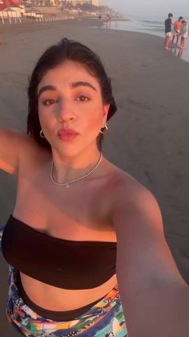 A fair-skinned woman with dark hair, wearing a black bandeau top and a colorful wrap, is on a beach at sunset. She's recording a selfie video, holding the camera up high. Other people are visible in the background, walking along the shoreline. The waves are gently rolling onto the shore. She pivots to the right and another woman with fair skin, wearing a light-colored top, blue shirt, and striped shorts smiles at the camera. The woman recording the video turns back to face the camera. Her expression is neutral. The light reflecting from the setting sun illuminates her face.