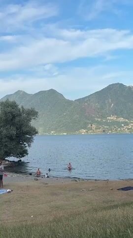 A scenic view of a lake with mountains in the background. On the grassy shore, several people are relaxing and swimming in the water. A large tree stands to the left, providing shade. The sky is blue with scattered clouds.