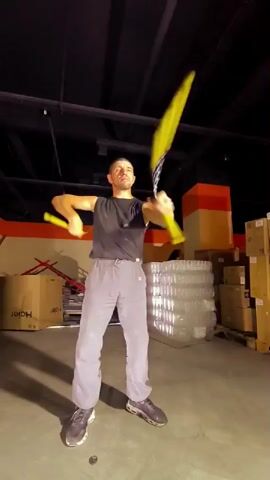 A man in a black tank top and grey sweatpants juggles two yellow poi in a dimly lit warehouse. The poi are blurred with motion, creating streaks of yellow light. The man's face is visible as he concentrates on his juggling. The background is filled with stacked boxes and plastic-wrapped items, suggesting a storage or distribution facility. The floor is concrete.