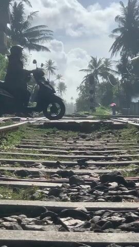 This vertical video shows a man on a scooter stopped on a set of railroad tracks. The tracks are lined with wooden sleepers and gravel. Palm trees and lush green vegetation flank the tracks, creating a tropical setting. The sky is cloudy with bright sunlight filtering through, suggesting it's daytime. In the distance, a person holding a pink umbrella walks along a path near some buildings. The atmosphere appears calm and perhaps a bit humid. The scene is set in Kodagoda, Sri Lanka. No text is visible in the video.