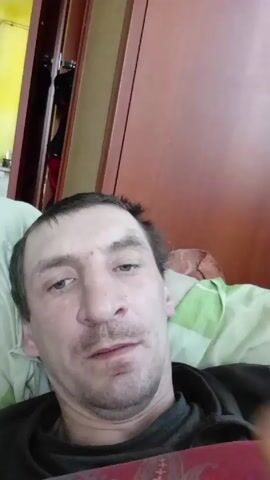 A man is lying down and touching his nose with his finger. He looks slightly annoyed or disgusted. He is lying on a patterned blanket in what appears to be a bedroom. A door is visible behind him, and a blurred image of a room with a stove can be seen in the background.