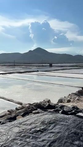 The video captures a wide shot of salt evaporation ponds under a bright, partly cloudy sky. The ponds are rectangular and appear to be filled with shallow water, reflecting the sky. In the foreground, there is a dark, textured surface, possibly a tarp, with a border of rough stones. In the mid-ground, more salt ponds are visible, separated by low, stone walls. In the background, rolling hills or mountains rise against the horizon, with a clear blue sky and scattered white clouds overhead. Power lines and poles are visible in the distance on the left side of the frame. The overall scene suggests an open, arid landscape utilized for salt production.