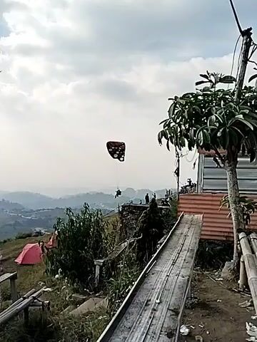 A butterfly-shaped paraglider is visible against a hazy sky, with a person suspended beneath it. The paraglider is flying over a hilly landscape with scattered buildings and vegetation. In the foreground, there is a tall tree, a pink tent, and a wooden structure. The scene appears to be a scenic viewpoint, possibly a tourist attraction.
