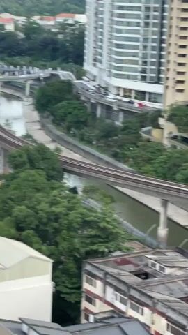 A high-angle aerial view pans across a city landscape. A river flows through the center of the frame, with elevated train tracks running alongside it on either side. Buildings of varying heights, including a tall, modern skyscraper, are visible in the background, interspersed with lush green trees. Several bridges cross the river, and traffic can be seen on a multi-lane road in the distance. The sky is overcast.