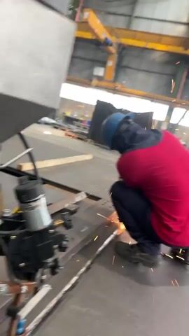 The video shows a person in a red shirt and blue helmet welding in a factory. Sparks are flying from the welding torch. Another person, wearing a grey shirt and gloves, is partially visible in the foreground, facing away from the camera. The environment appears to be a large industrial space with a crane visible in the background. The video is shaky and blurry at times.
