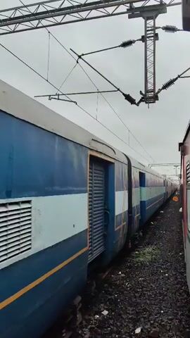 The video shows a train on parallel tracks under a cloudy sky. The train is blue with light blue and yellow stripes. The side of the train has windows and what appear to be ventilation grates. In the foreground, there are train tracks and gravel. The camera is moving, showing a side view of the train as it passes by. There are yellow circular signs visible on the side of the tracks. The train appears to be stationary.