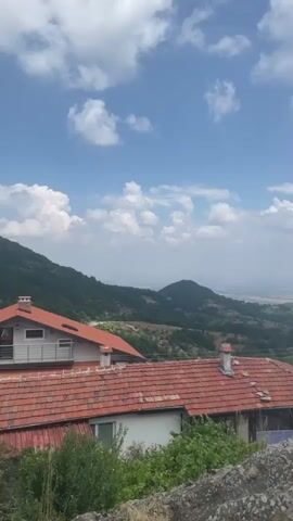 The video pans across a rural mountainous landscape on a cloudy day. In the foreground, there are rooftops of houses with red tile roofs, interspersed with green vegetation and rocks. In the middle ground, rolling green hills and more distant mountains are visible. The background shows a hazy, flat expanse that extends to the horizon. A black metal pole with decorative elements and writing in Cyrillic script is visible in the center of the frame. The sky is blue with scattered white clouds.