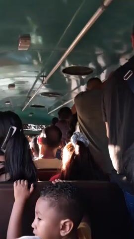 The video depicts the interior of a crowded bus. The roof is green and has metal handrails and vents. Passengers are standing and sitting. Some are holding onto the rails.  A man in a black shirt and cap is standing near the camera.  There is a small child in the foreground with a red balloon.  The overall scene is one of public transportation with many people in a confined space.