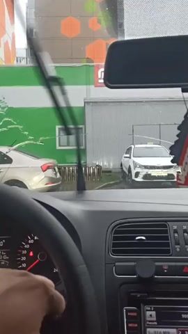 The camera is inside a car on a rainy day, with raindrops visible on the windshield. Through the windshield, we see a light gold car and a white Kia parked in front of a building with green and white walls, decorated with orange hexagons. The camera's focus is primarily on the exterior scene, with the car's interior elements—steering wheel, dashboard, rearview mirror—partially visible and framing the view. A hand can be seen holding the steering wheel. The wipers appear to be off.
