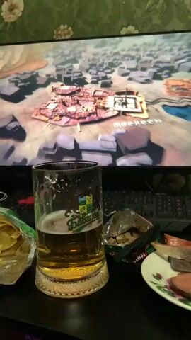 A close-up, eye-level shot shows a mug of beer in the foreground, with a computer screen displaying a video in the background. The beer mug is half-full and appears to be golden in color, with a frothy head. To the right of the mug, a plate holds several slices of what appears to be cured meat. To the left of the mug, a bag of potato chips is visible. The computer screen displays a scene from what looks like a video game or a television show, featuring an aerial view of a city or settlement. Text overlays on the screen indicate credits, with names like "CAROLYN STRAUSS," "DAVID BENIOFF," and "D.B. WEISS" visible. The overall environment appears to be a dimly lit room, likely a desk or table, with a computer keyboard visible beneath the screen. The wallpaper behind the screen has a floral pattern. The video plays for a few seconds, showing subtle animations or transitions on the screen.