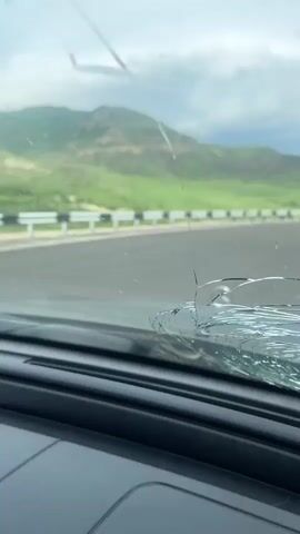 The video is filmed from the driver's perspective inside a vehicle, identified by a Mercedes-Benz hood ornament prominently displayed on the dashboard. A noticeable crack or significant dirt accumulation is present on the right side of the windshield.

The vehicle is on an asphalt road that curves slightly to the right. To the right of the road, a white guardrail runs alongside the shoulder. The background features lush green, rolling hills and mountains, characteristic of a rural or semi-mountainous landscape, consistent with the given location context of Vardashat, Armenia. The sky is heavily overcast with white and gray clouds, indicating daytime but a subdued light.

On the right side of the road, beyond the guardrail, a group of approximately 5-7 people is gathered. Among them, a brown horse is visible, being held or attended to by one person. Further down the road, a white police vehicle with flashing blue lights can be seen, and at least one individual appears to be a police officer, dressed in a dark uniform. The people seem to be observing or interacting, possibly in response to a roadside stop or an incident involving the horse. The overall impression is a roadside gathering or official stop in a scenic, mountainous region. No discernible text is visible in the scene other than the Mercedes-Benz emblem.