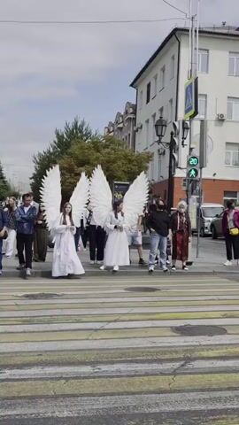 A group of people are walking across a crosswalk on a paved street. In the foreground, two individuals with large white angel wings are walking together. They are both wearing white robes. To the right of them, a woman in a dark shirt and jeans is walking. Behind them, other pedestrians are visible, including individuals in casual clothing. A traffic light with a green signal is visible on a pole to the right of the crosswalk. Buildings line the street in the background. The sky is overcast.