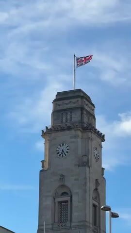 A tall, light-colored stone clock tower stands prominently against a bright blue sky with scattered white clouds. A Union Jack flag, displaying the colors of the United Kingdom, flies from a flagpole at the very top of the tower, gently waving in the breeze.

The clock tower has multiple levels, with the main clock faces situated on the upper section. There are two visible clock faces, one on the left and one on the right, both appearing to show a time around 2:45 PM. Below the clock faces, there are decorative architectural details and arched windows with patterned grilles. The overall design suggests a classical or civic building style.

In the bottom right corner, a street light fixture is partially visible, indicating an urban setting. The video begins with the sound of a passing vehicle, further reinforcing the urban environment of Barnsley, United Kingdom. The scene is bright and clear, suggesting a sunny day.