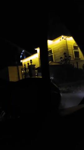 A dark, nighttime video shows a yellow house with illuminated windows and exterior lighting. A black metal gate is visible in front of the house. A portion of a car, including a wheel, is seen in the foreground. The video appears to be filmed from inside a vehicle, looking out through a side mirror. There are no people visible in the video.