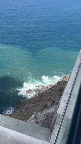 The video shows a high-angle view of a rocky coastline meeting the ocean. The camera pans from a concrete ledge with a glass railing to reveal the coastline. The ocean transitions from a dark blue further out to a lighter turquoise closer to the shore. Waves crash against the rocky cliffs. The wind is strong. The sky is partially cloudy. The coastline extends into the distance with multiple points jutting into the ocean.