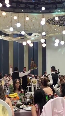 The video shows a scene from a lively party.  Numerous people sit at tables adorned with food and drinks, in what appears to be a banquet hall. The hall is well-lit with numerous round, pendant lights hanging from the ceiling, along with decorative white ribbons. A waiter in a black vest and white shirt is walking through the tables, holding high a platter of food, possibly meat skewers, arranged around a decorative wooden figure of a man on horseback.  He is being closely followed by another waiter carrying another platter. The atmosphere appears festive.