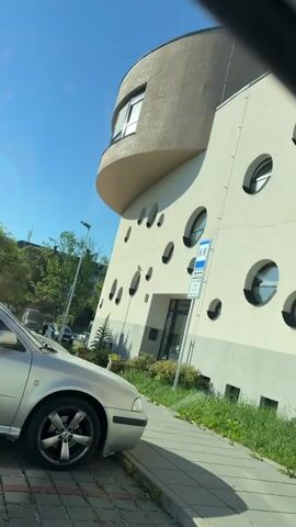 The video captures a sunlit daytime scene in Olomouc, Czechia. A silver car is parked on a paved area next to a sidewalk with overgrown grass. Behind the car, a distinctive building with a curved, beige upper section and a white, cream-colored facade is visible. The building's facade is adorned with numerous irregularly spaced, oval-shaped windows, some of which are deep-set and create dramatic shadows.

A blue and white parking sign with text in a foreign language is affixed to the building. The text is partially visible and appears to be "PLATÍ V DOBĚ PO-PA 6,30-19,30 MAX. 15MIN". The building also has a darker section near the entrance, suggesting a doorway or shopfront with a sign that is difficult to read, but it seems to include the word "Waldorf".

The sky is a clear, bright blue, indicating fair weather. Lush green trees line the background, suggesting a verdant urban environment. The overall mood is peaceful and ordinary, depicting a typical street scene in a European city on a sunny day. The focus is on the unique architecture of the building and its unusual windows, as well as the parked car and the natural elements of the surroundings.