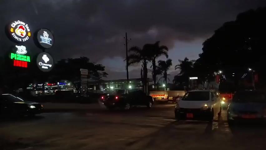 The scene is an outdoor setting at night. The sky is dark with some clouds. In the foreground, there are several cars parked or slowly moving on a road. In the background, there is a building with several illuminated signs for various food establishments, including "Chicken Inn", "Pizza Inn", and "Creamy Inn". There are also several palm trees and other trees silhouetted against the dark sky. The overall scene depicts a typical night scene in a commercial area.