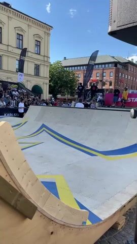 The video shows a skateboarding event in Karlstad, Sweden. The scene is outdoors on a sunny day with blue skies. A skate ramp, made of wood with blue and yellow painted designs, is the central focus.

Several people are present, including skateboarders and spectators. Some skateboarders are gathered at the top of the ramp, while others are seated or standing around the event area. Spectators are watching from a tiered seating area behind the ramp.

Visible text includes "SM VECKANI" and "VÄRMLAND" on a black banner, suggesting a national competition or event. Another banner has "KARLSTAD" printed on it. The architecture in the background indicates an urban setting, with buildings of varying styles. The overall atmosphere appears to be lively and festive, typical of an outdoor sporting event. The time of day is likely midday, given the bright sunlight and clear skies.
