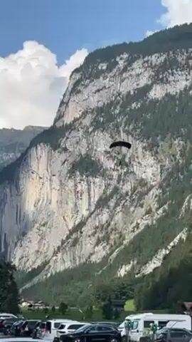 The video shows a skydiver parachuting against the backdrop of a tall, rocky mountain. The mountain has streaks of gray rock interspersed with green trees.  The sky is blue with some fluffy white clouds. The skydiver, under a dark canopy, descends towards the valley floor, where a parking lot with cars and campers is visible. Some green structures are also seen in the parking area. Voices in a language other than English can be heard.