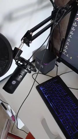 The video shows a close-up, top-down view of a small section of a desk, focused on computer and audio equipment. A black microphone is mounted on a black articulated arm with a pop filter. A small, dark gray speaker cube is behind the microphone. To the right, there is a black keyboard with blue backlighting and part of a computer monitor displaying various icons. Numerous cables are visible, connecting the devices. The desk surface is white. Music is playing in the background.