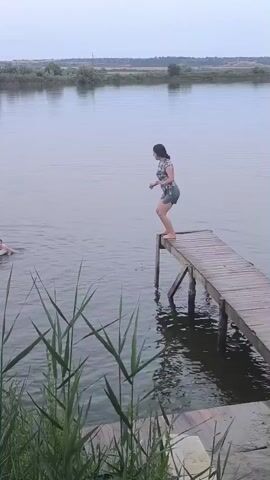 A young person jumps off a wooden dock into a body of water. Another person is already in the water, to the left of the frame. The water is calm, with reeds growing along the bank in the foreground. The background consists of a distant treeline and a hazy sky. The jump is captured in a series of stills, showing the motion of the person from the moment of jumping to their entry into the water, creating a splash. The person in the water to the left appears to be watching the jump.